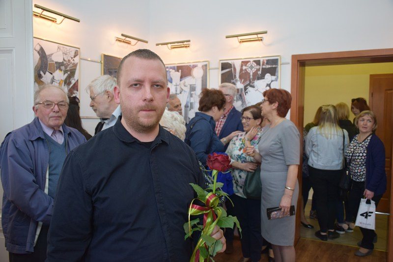 Maciej Małecki, the artist, standing at a gallery opening holding a single red rose, framed works on the wall behind him.